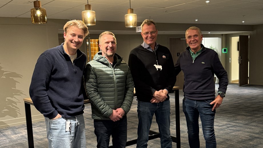 Programledere og gjester står oppstilt i et stort rom med taklamper i bakgrunnen. Alle smiler og ser mot kamera.