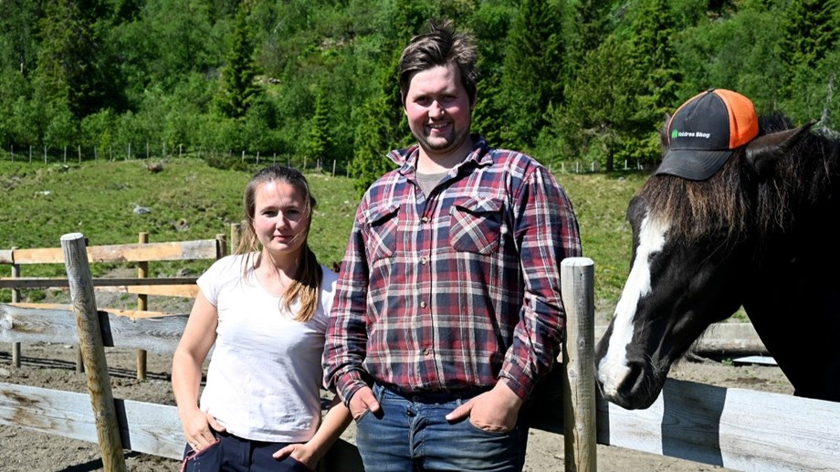 Thea og Jørgen står ute foran et gjerde med en hest som står bak gjerdet. De ser mot kamera og hesten har på seg en caps.