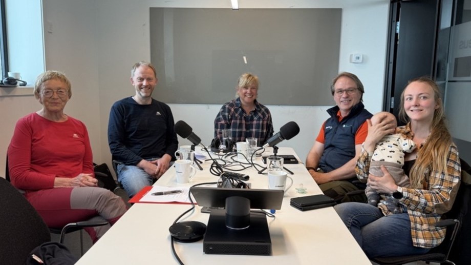 Gjester og programledere sitter rundt et bord og smiler mot kamera. Mikrofoner på bordet.