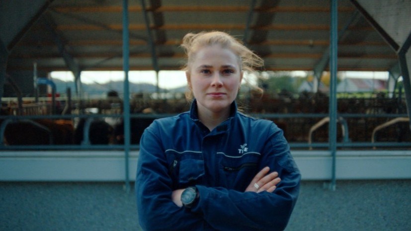 Ung kvinnelig bonde står ute foran fjøset med kyr. Hun holder armene i kors og ser mot kamera.