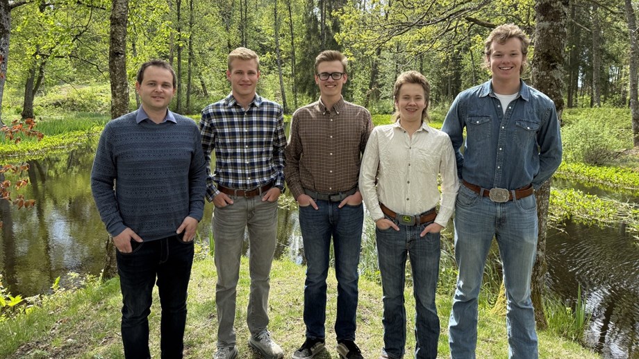 Læreren og elevene står oppstilt på rekke utendørs. I bakgrunnen vises et lite vann og en skog.