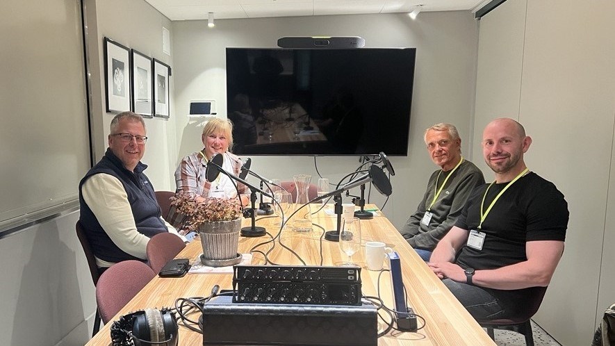 Programledere og gjester sitter rundt bordet i studio med mikrofoner foran seg på bordet.